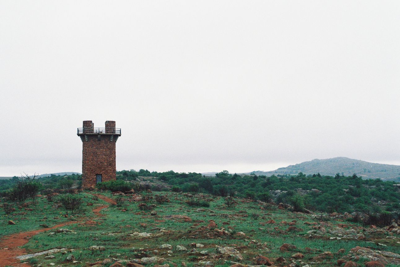 A red stone tower stands off-center on a red dirt and green scrub landscape against a grey sky.