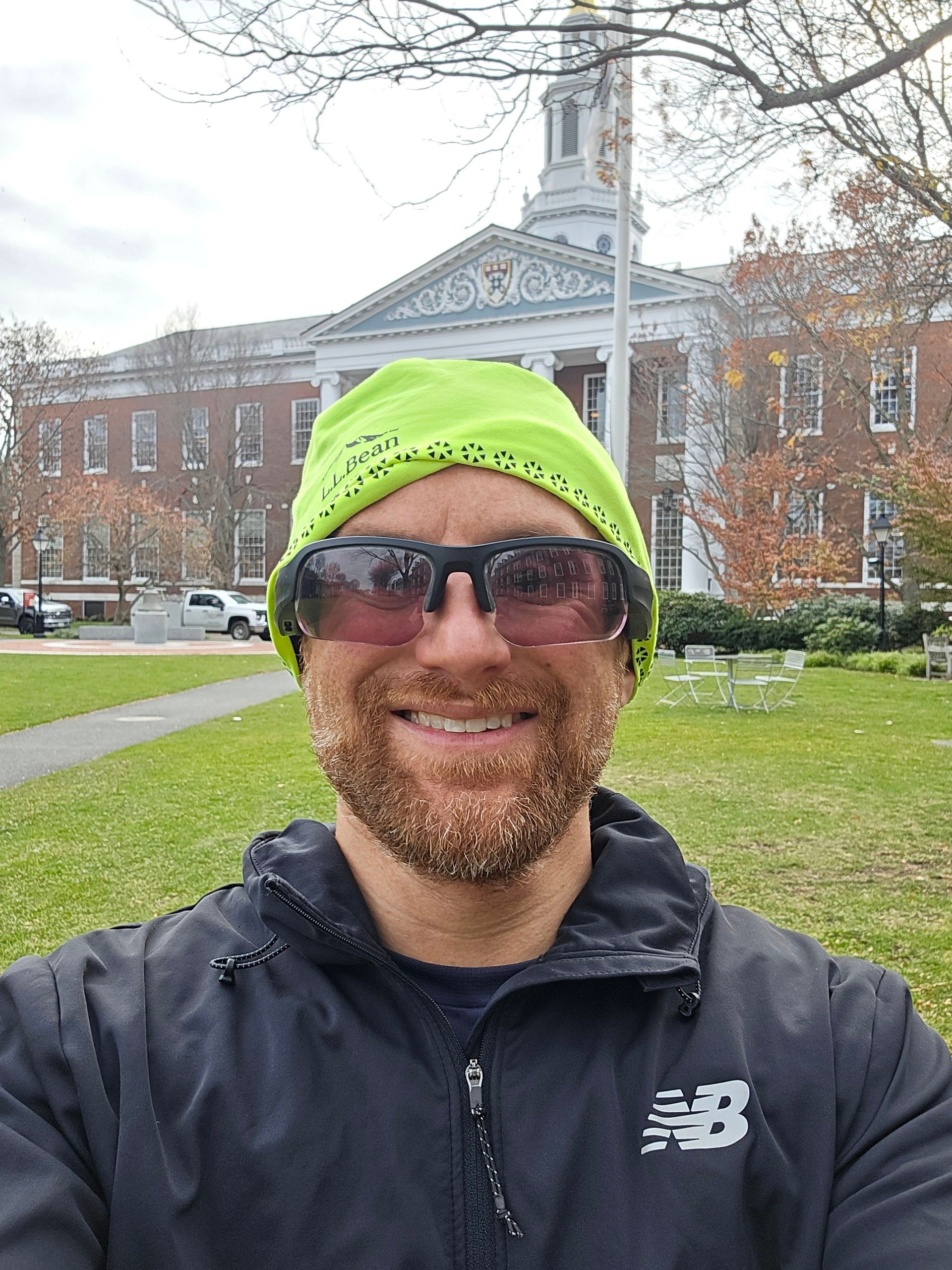 A selfie of me in front of Mellon Hall at HBS on a bright day. I’m a middle-aged white man with a red beard flecked with white. I’m wearing a neon green LL Bean beanie, black sunglasses, and a black New Balance running jacket over a dark blue running shirt.