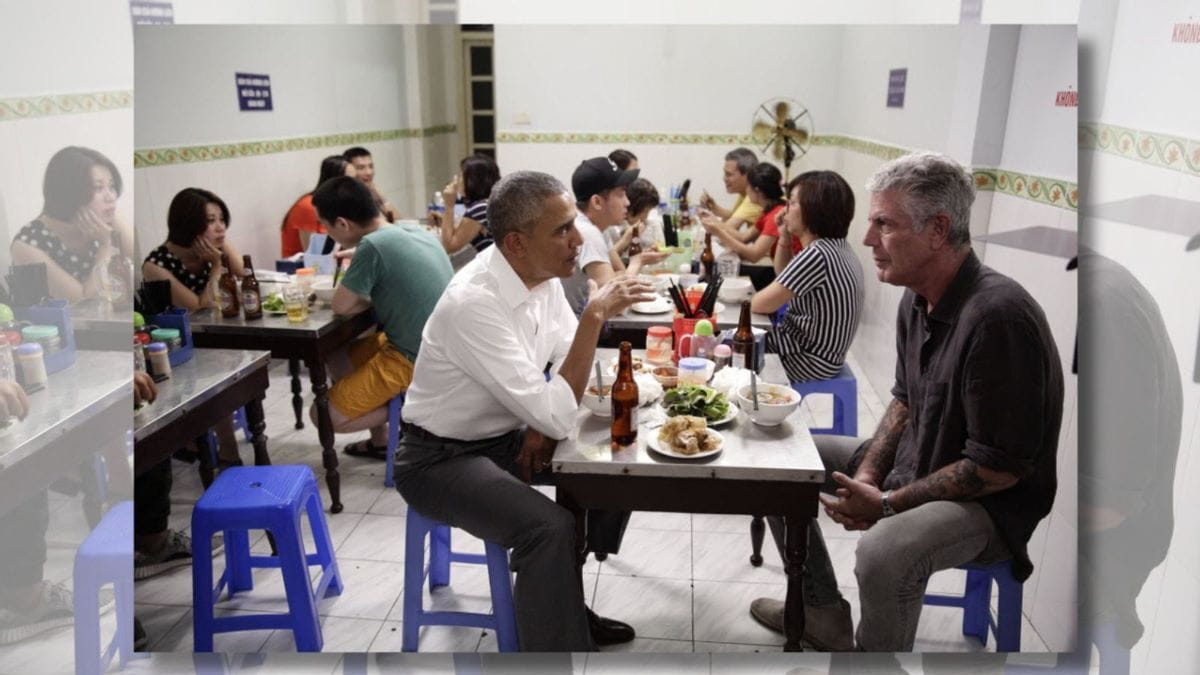 A Black man sits across from a white man at a table with food and beers. Several people sit at other tables around them.