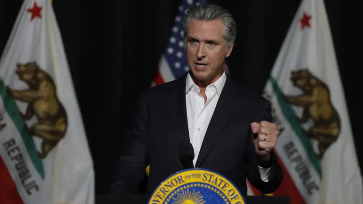 A white man wearing a black suit stands at a podium while speaking. His right hand is clenched in a fist.