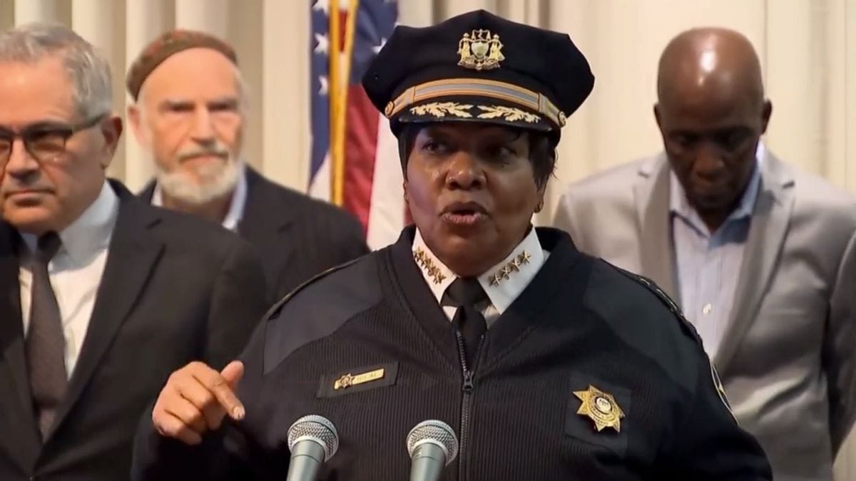 A Black woman stands at a podium speaking into two microphones. Several people can be seen standing behind her. 