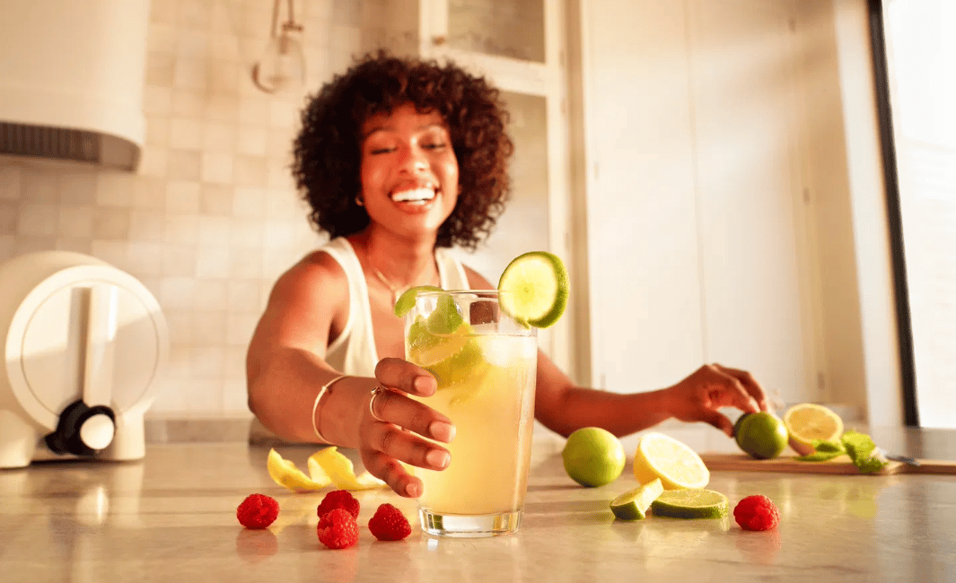 A woman smiling and reaching for a glass of kombucha.