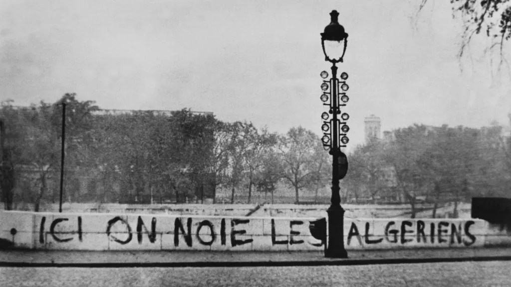 A photo of a riverbank and lamppost. Painted on the low wall along the bank are the words "ICI ON NOIE LES ALGERIENS." 