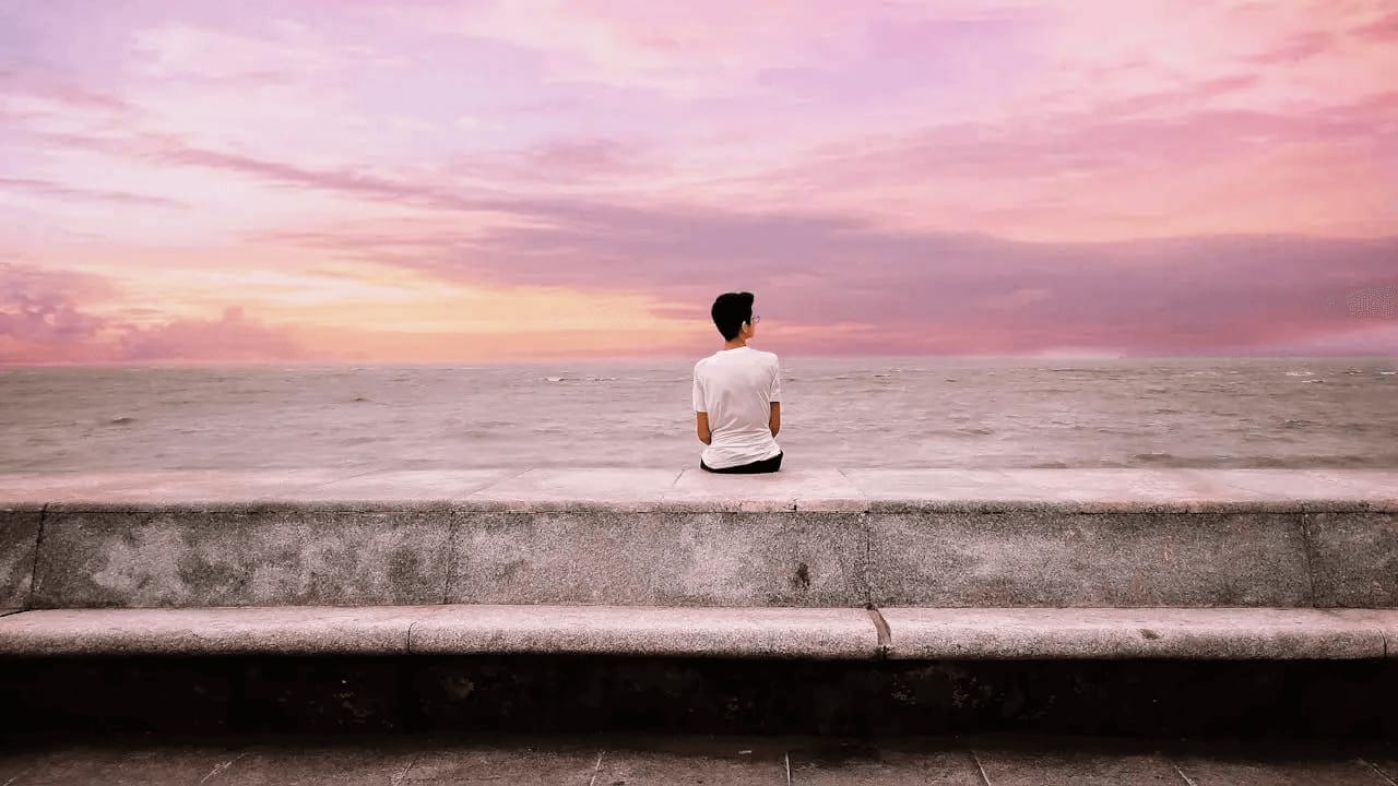 man sitting on a break water looking out over the water towards a pink sunset
