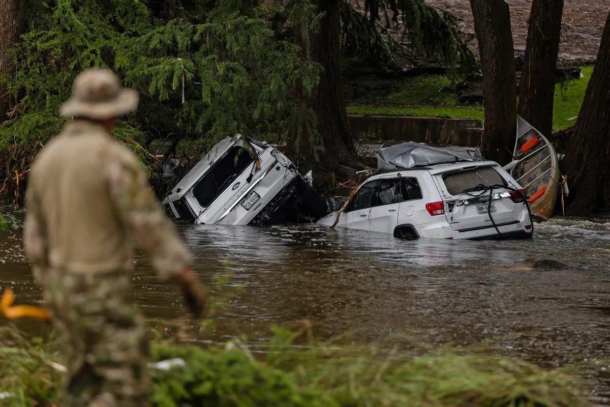 A person in military-style clothing stands in the foreground, observing a flooded area where two heavily damaged SUVs are partially submerged and wedged against trees. One vehicle is tilted on its side and a small overturned boat is also caught among the trees. The scene shows the aftermath of a severe flood, with fast-moving water and debris scattered around.