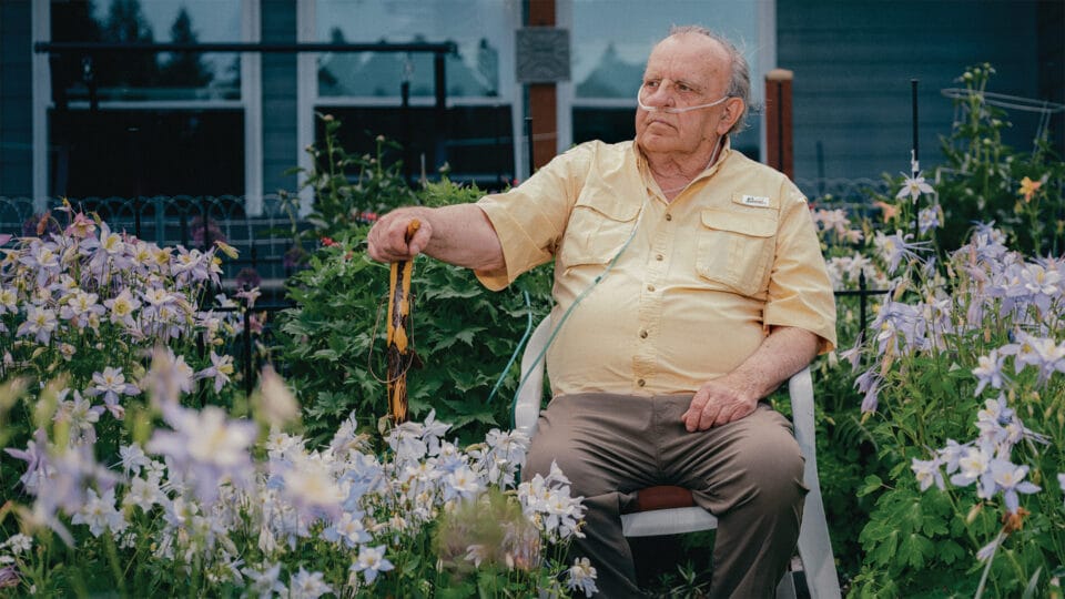 “Elderly man with an oxygen tube sits on a white chair in a garden full of blooming flowers, holding a cane and gazing thoughtfully into the distance.”