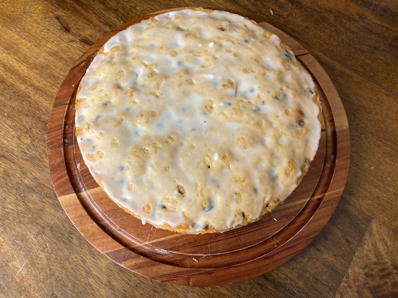 A round cake drizzled with a thin lemon glaze and studded with specks of rosemary sits on a wooden cake board.
