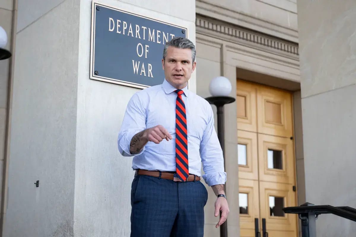 A white man stands in front of a building sign that says DEPARTMENT OF WAR.