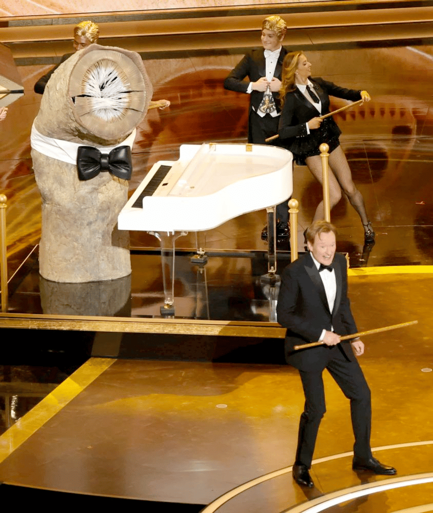 A crude sandworm costume stands at a white piano whle Conan O'Brien and others sing and dance