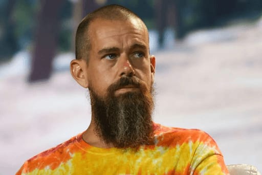 Jack Dorsey with a long beard and shaved head, wearing a tie-dye shirt, seated with a serious expression against a blurred outdoor background.