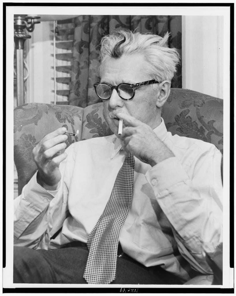 black-and-white photo of a man with white hair and a black streak in thick glasses, sitting on an upholstered chair lighting a cigarette with a match and looking down at the flame