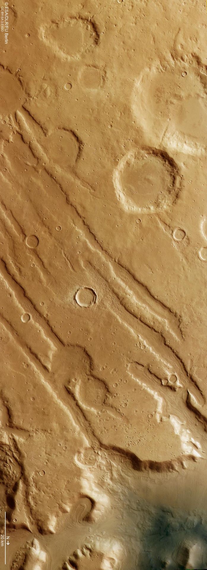 As we move northward from Mars’s equator, we encounter Coloe Fossae—a region etched with narrow grooves, deep valleys, scattered impact craters, and evidence of a past ice age. This image, captured by ESA’s Mars Express High Resolution Stereo Camera on 19 October 2024 (orbit 26257), combines data from the nadir (the field of view aligned perpendicular to the surface of Mars) and color channels to offer a detailed view of the terrain. North is to the right, and the image resolution is roughly 16 meters per pixel, centered near 39°N/54°E.