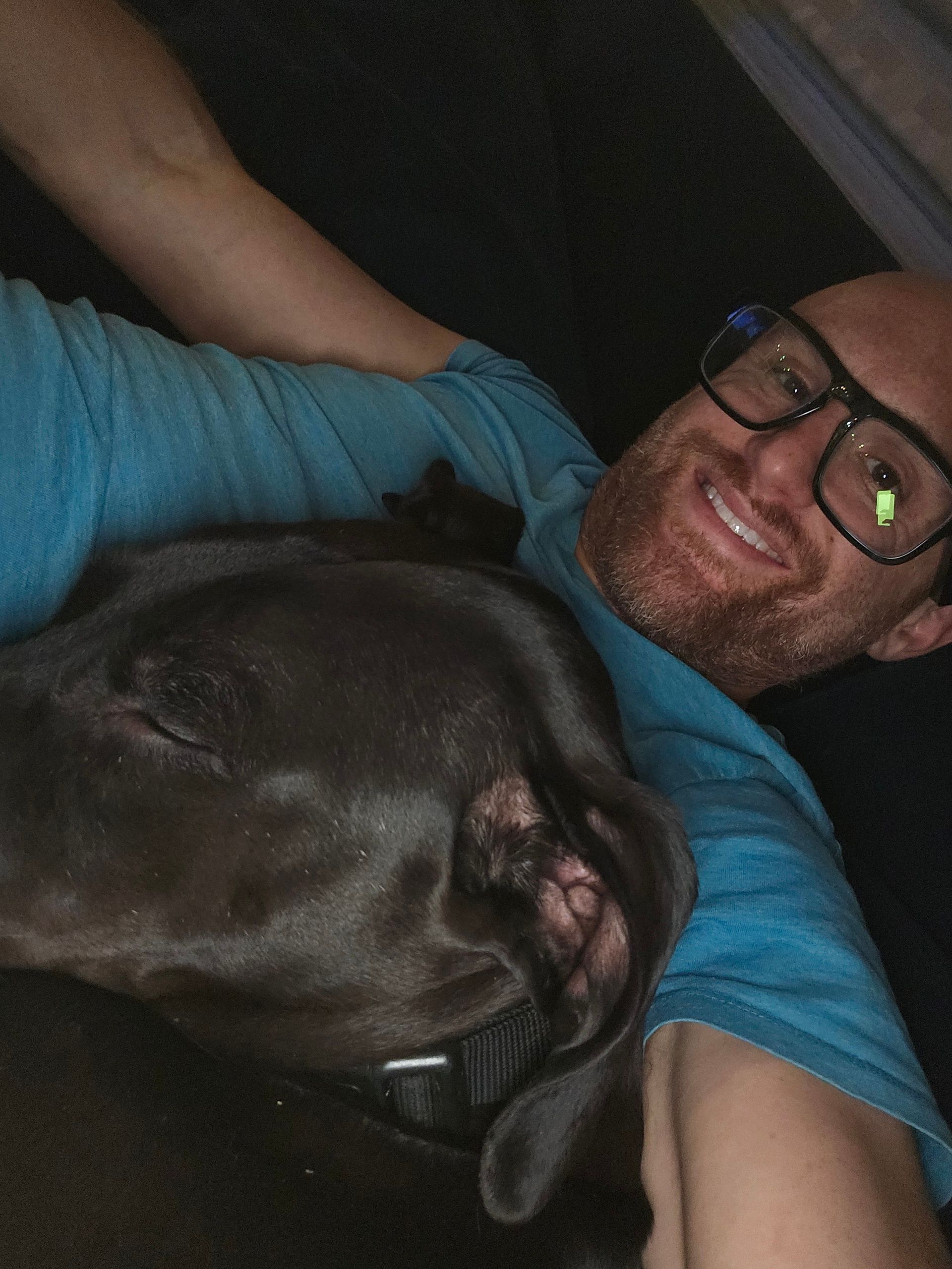 A selfie of me on a couch with my medium-sized black dog sleeping with his head laying on my chest. I'm a bald, middle-aged, white man with a red beard flecked with white. I'm wearing glasses with thick black rims and a blue t-shirt.