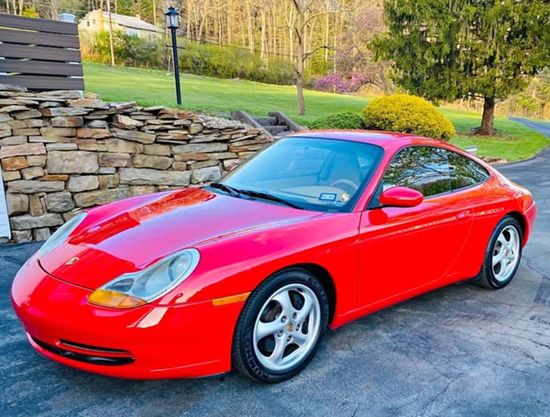 Front three-quarter view of a red 1999 Porsche 911 parked in a residential driveway with a stone wall background