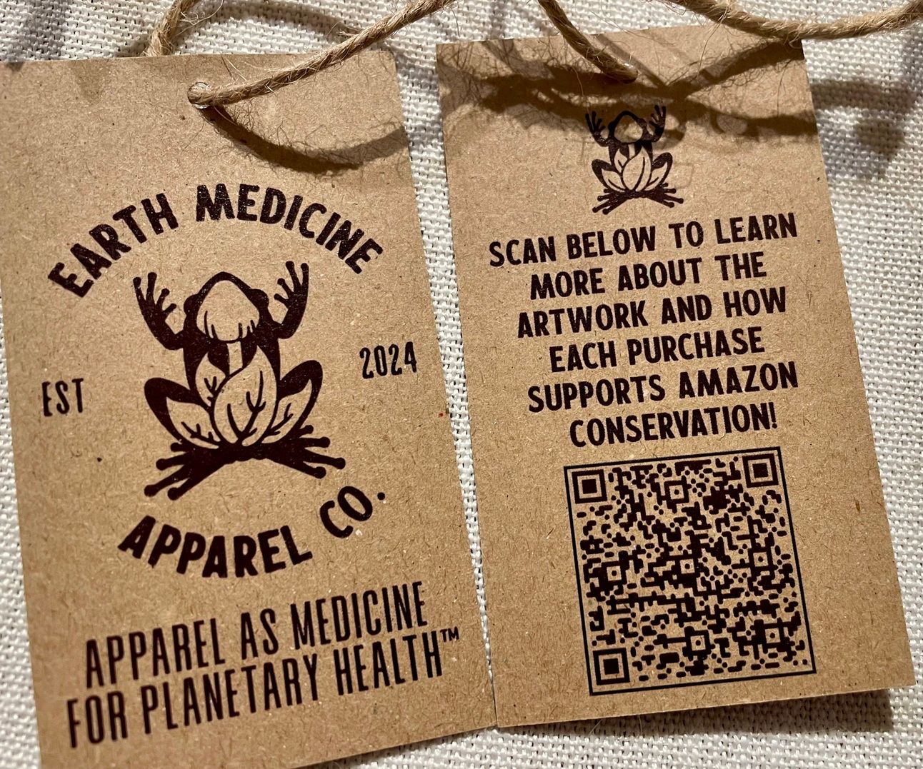 Front and back of kraft paper hangtags with dark purper ink, bearing company logo of a frog with a mushroom an leaves inside its body. Back reads "Scan below to learn more about the artwork and how each purchase supports Amazon conservation!" with a QR code.