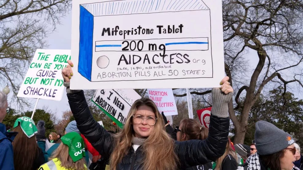 A protester holds a large hand-drawn sign depicting a box labeled “Mifepristone Tablet 200 mg” with the words “Aid Access—safe, legal, affordable—abortion pills all 50 states.” Other demonstrators and signs supporting abortion rights are visible in the background.