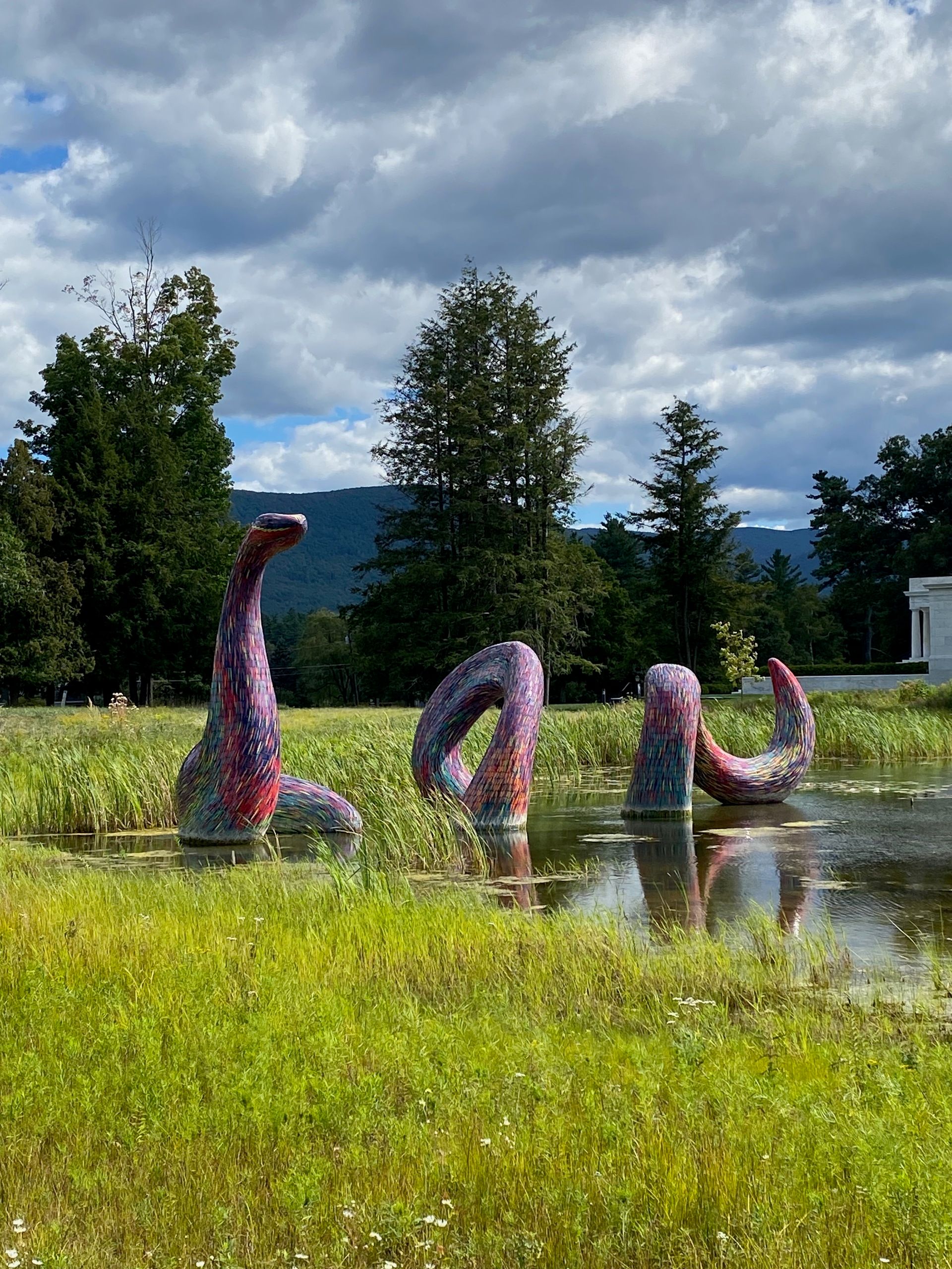 A large colorful statue of a serpent rising out of a small pond in front of trees and mountains.