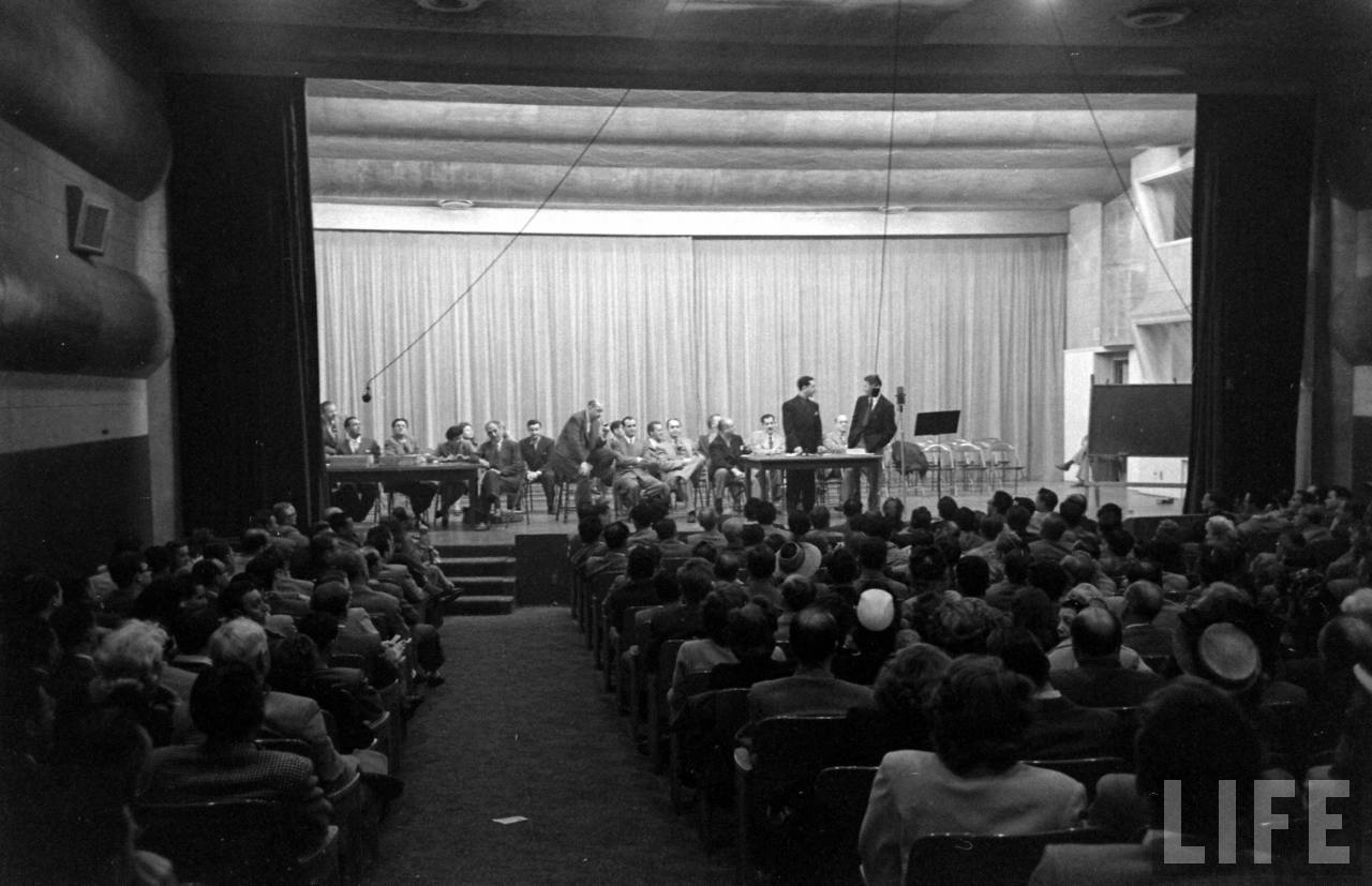 black and white photo of a crowd in a small theater facing a stage with several speakers, including two men standing befor microphones at a table.