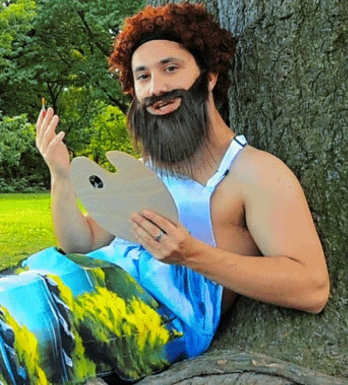 A man with a red curly wig and fake beard, dressed in a colorful artistic outfit, sits against a tree in a green park. He holds a painter's palette in one hand and gestures with the other, appearing to be in the middle of creating art. The scene is vibrant and playful.