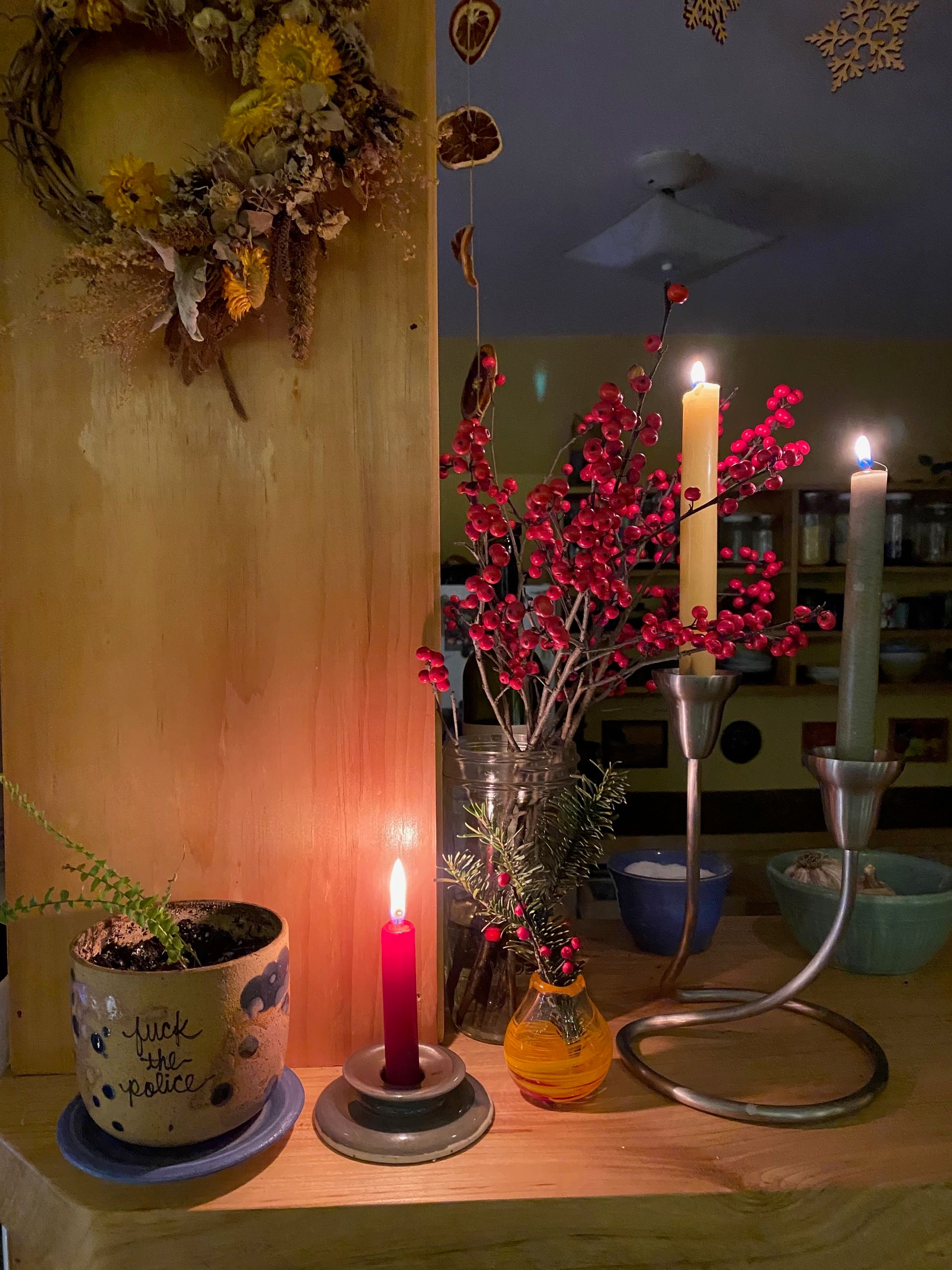 The corner of a wooden kitchen counter with two tall candles, a vase of winterberry, a small candle, and a tiny fern in a planter that says ‘Fuck the Police’.