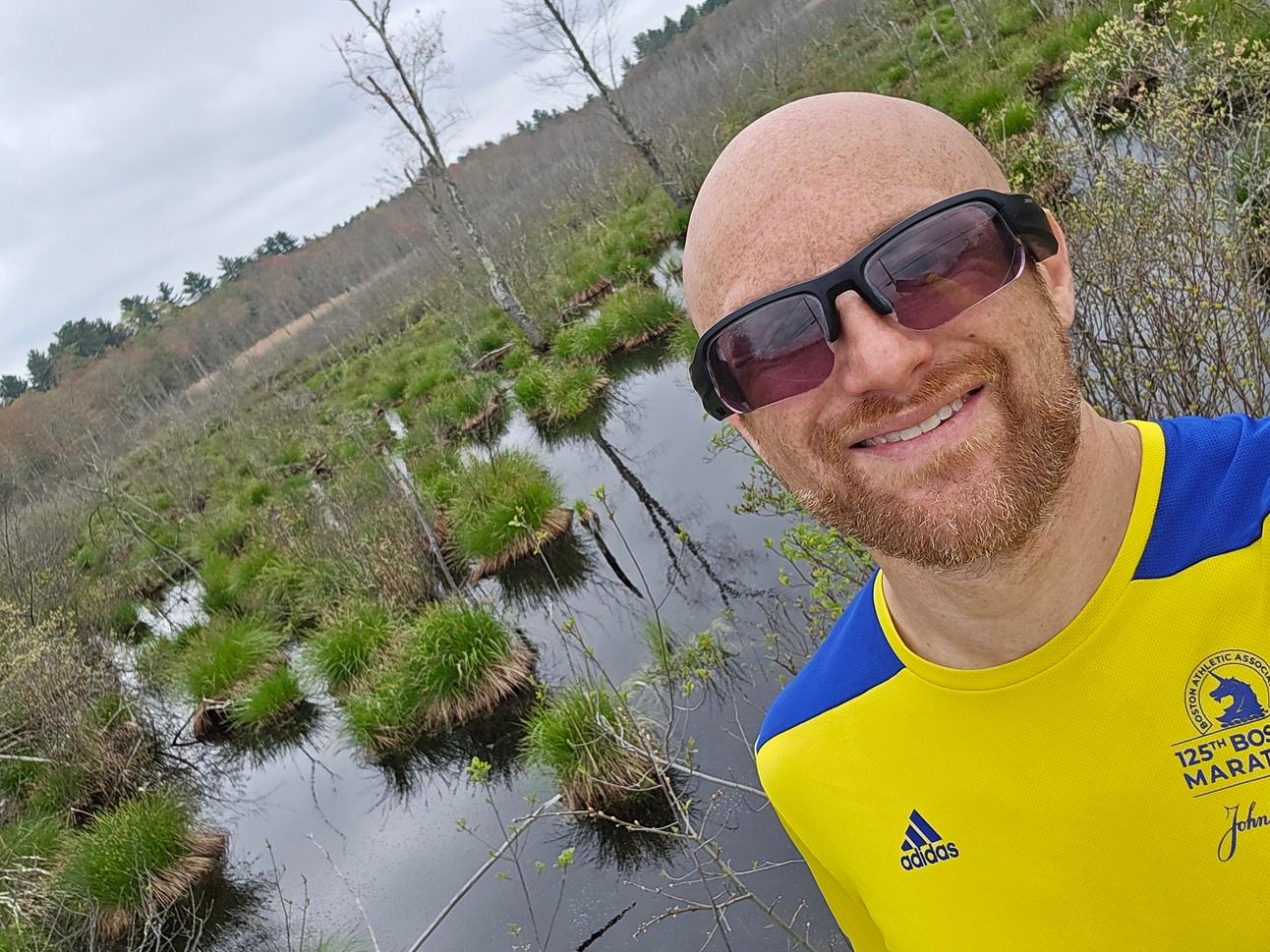 A selfie of me in front of a swamp on a cloudy day. Tufts of grass grow on individual clumps out of the water, with a few bare trees scattered throughout. Beyond is dense forest. I'm a bald, middle-aged, white man with a red beard flecked with white. I'm wearing black sunglasses and a yellow and blue 125th Boston Marathon adidas long-sleeved running shirt.
