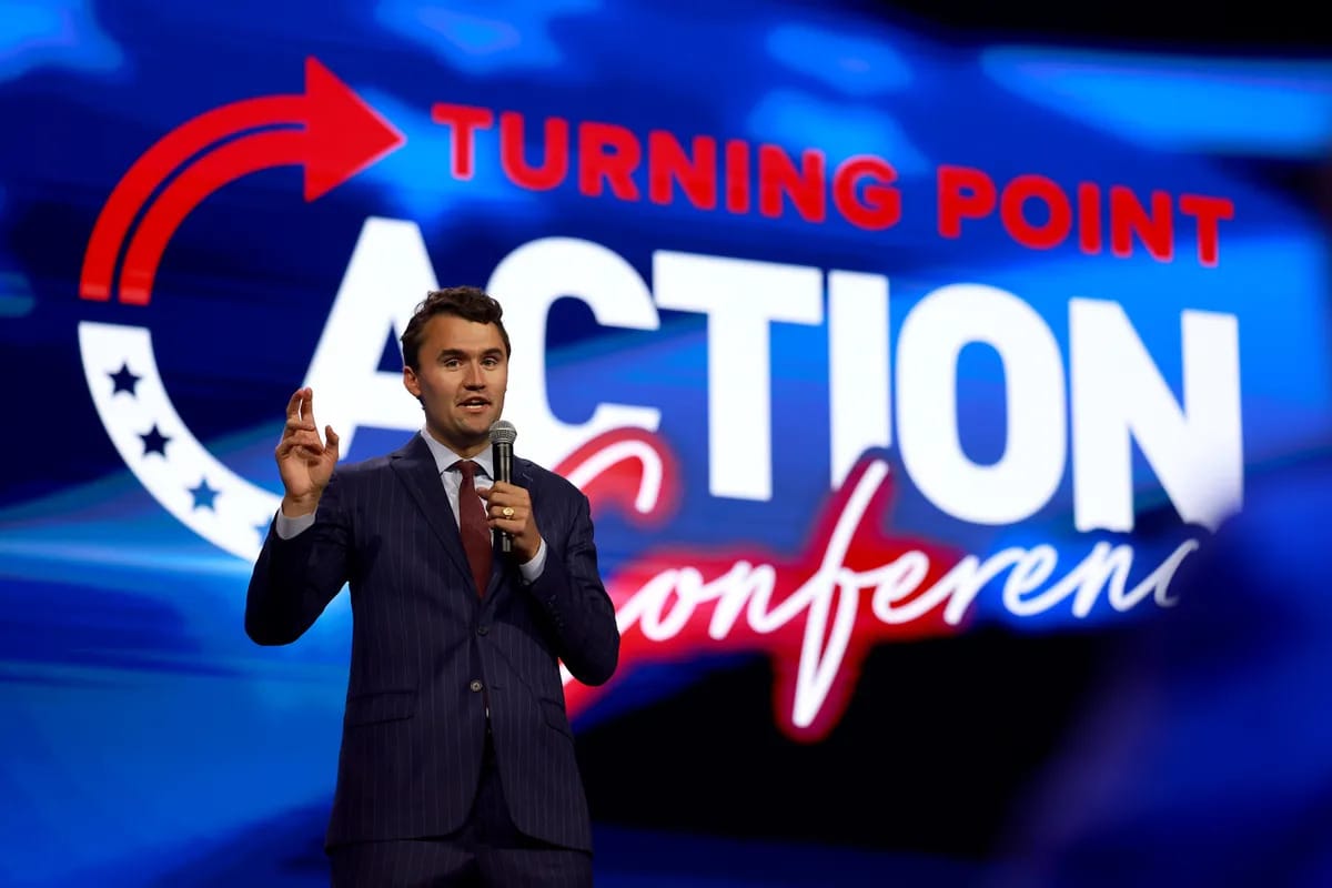 A white man holding a microphone talks in front of a sign that says TURNING POINT ACTUIN CONFERENCE.