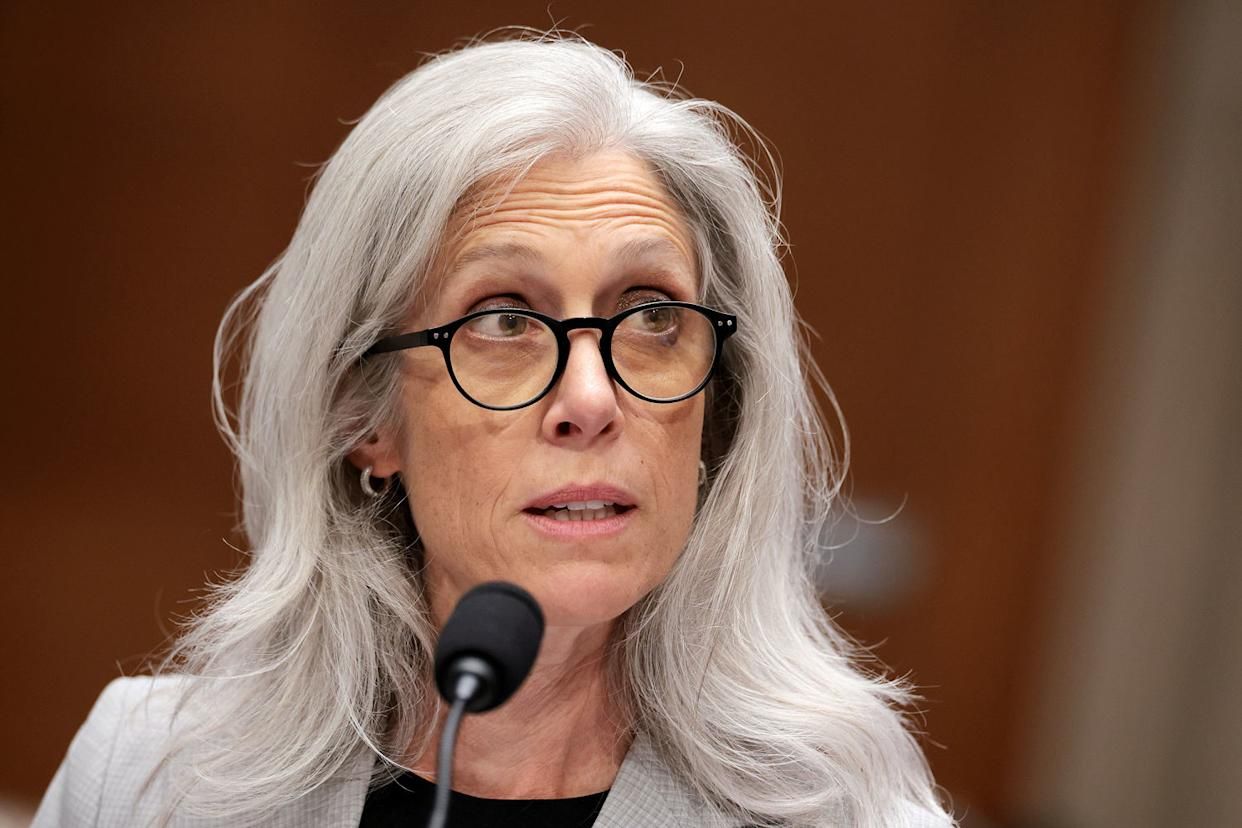 Woman with long gray hair and black round glasses speaking into a microphone at what appears to be a formal event or hearing.