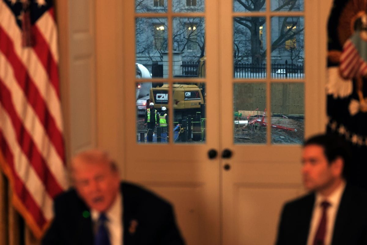 An image focuses on construction workers that can be seen working through a doorframe. Inside the room, two men wearing suits can be seen out of focus.