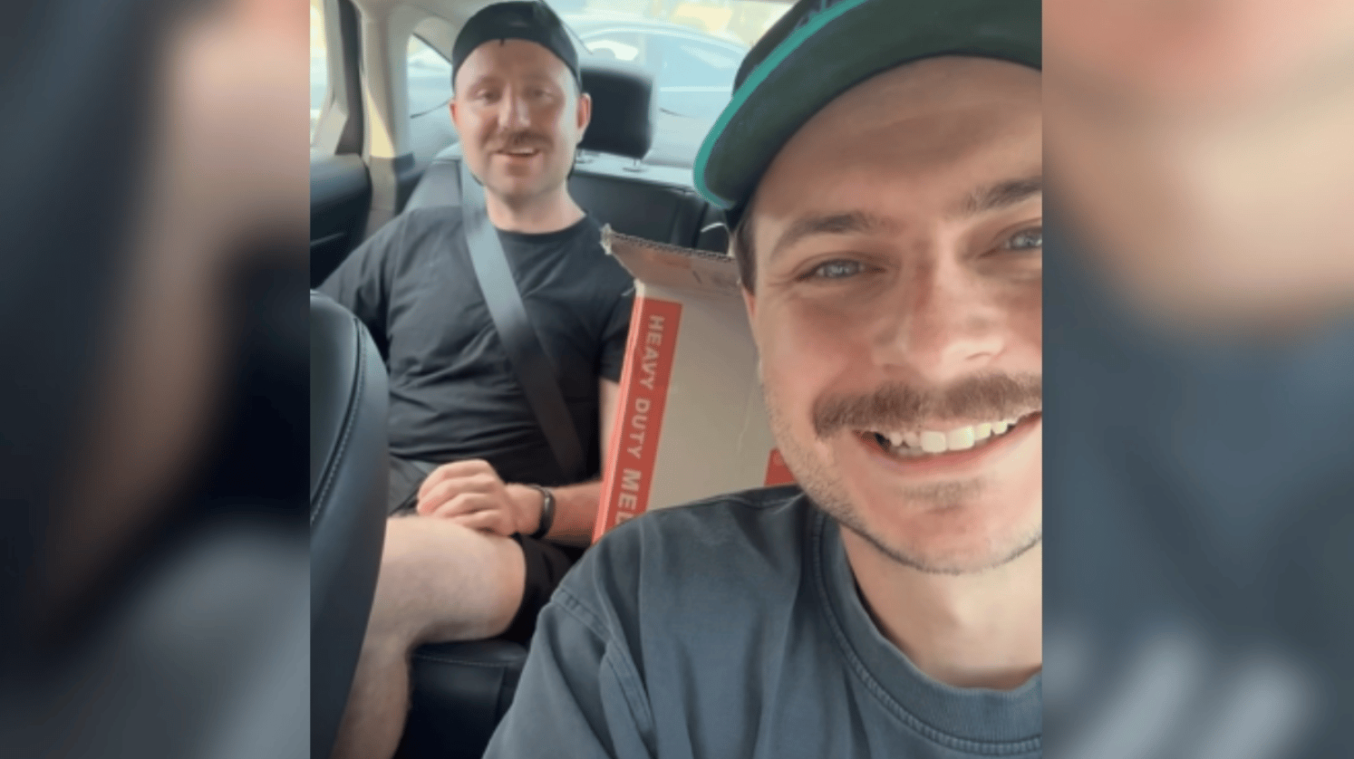 A man, who is holding the camera, smiles wide from the driver seat of the parked car. Another man sits in a backseat, also smiling