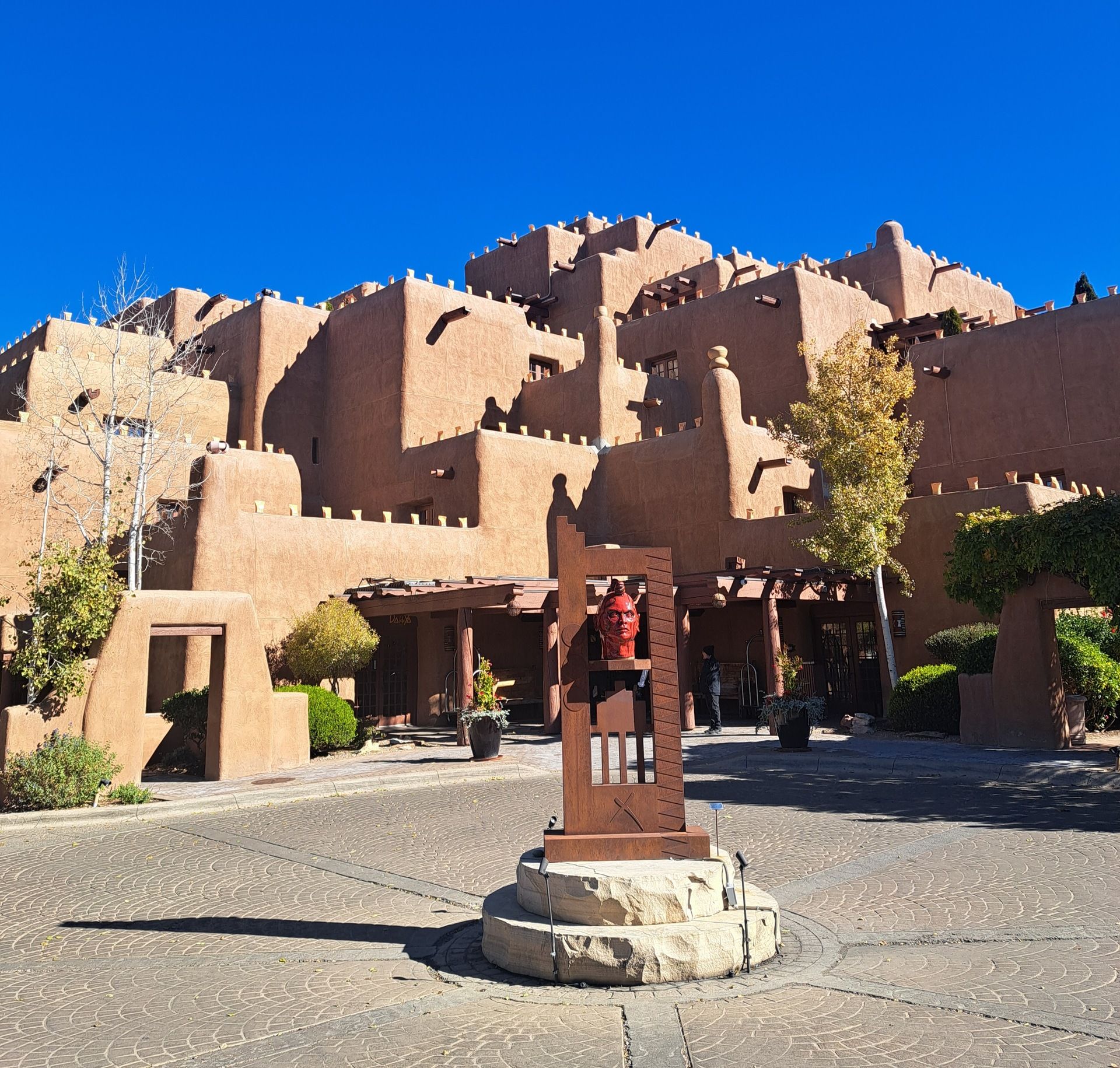 A photo of the Inn and Spa at Loretto in Santa Fe, New Mexico, featuring its distinctive adobe-style architecture with multiple terraced levels and rounded edges. The tan stucco building stands against a vivid blue sky. In the foreground, a striking red sculpture of a human face is mounted on a wooden frame in the circular driveway, surrounded by desert landscaping and potted plants.