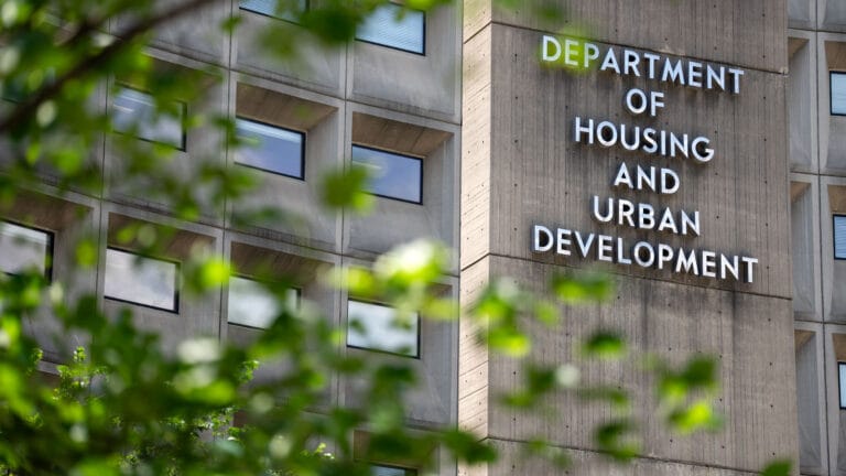 "Exterior of the U.S. Department of Housing and Urban Development (HUD) building, with its name displayed on a concrete facade surrounded by leafy green branches in the foreground."