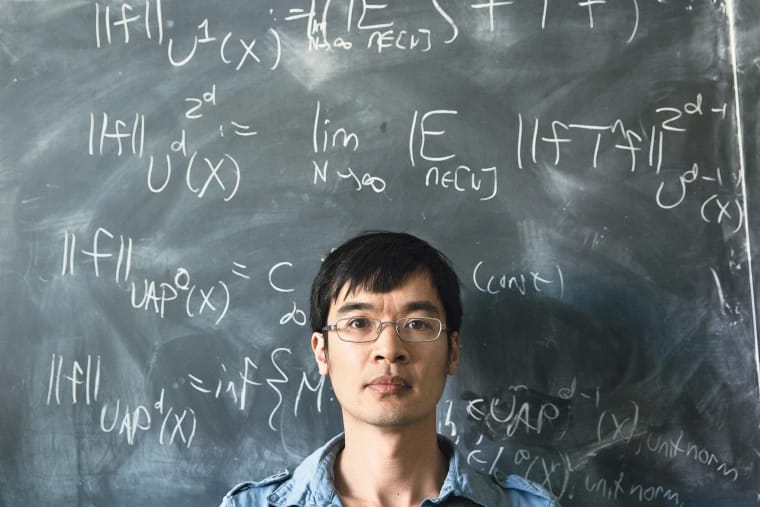 “A person standing in front of a chalkboard covered with complex mathematical formulas, wearing glasses and looking directly at the camera.”