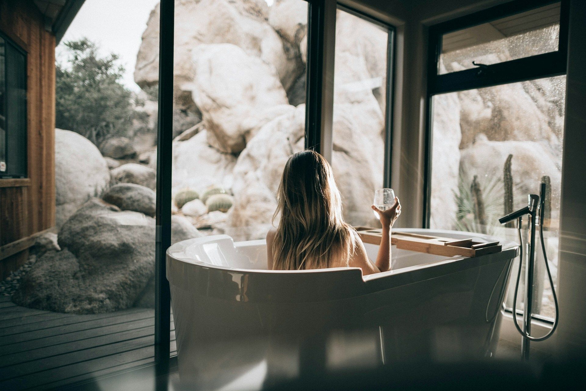 Woman relaxing in a white bathtub holding a glass of water or wine, looking out large windows toward desert rocks and cacti.