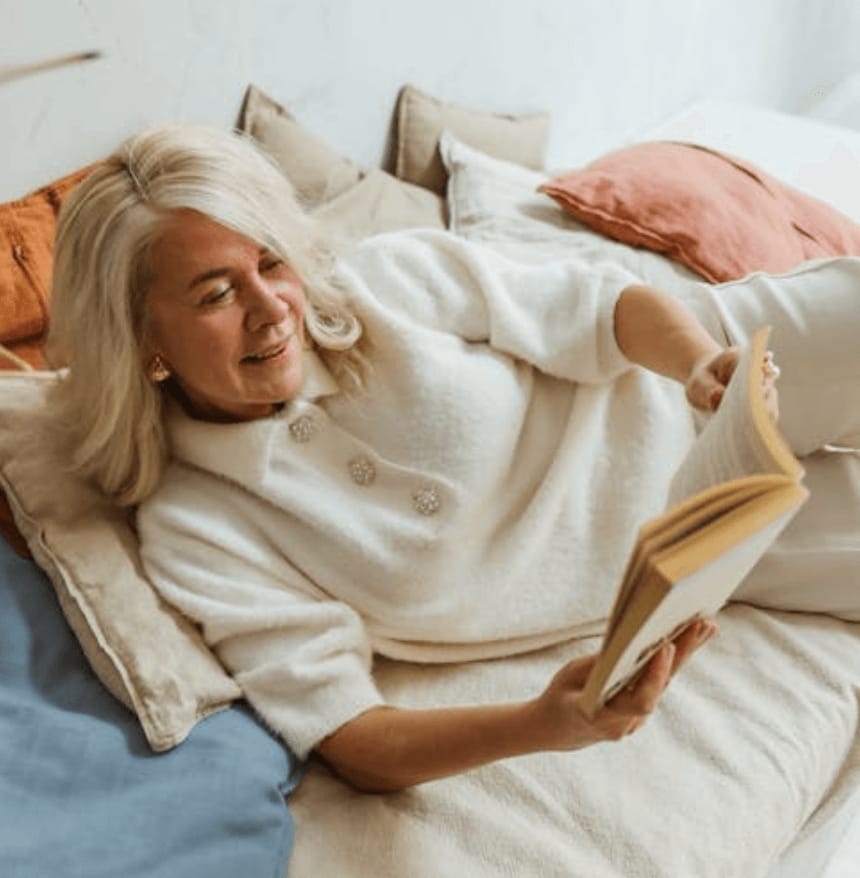 Older woman relaxing on a heap of pillows reading a book