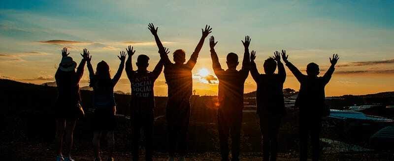 A group of friends silhouetted against the sunset with their hands raised — symbolizing unity, support, and collective joy.