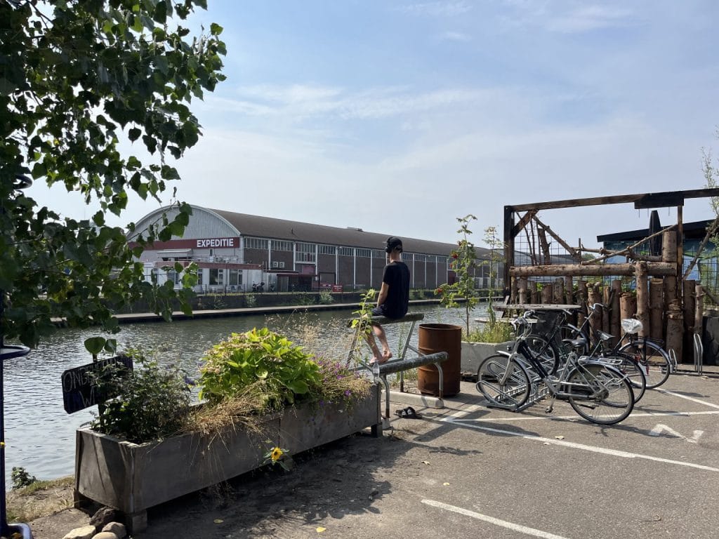 Zicht op een industriële kade aan het water met een groot gebouw waarop "Expeditie" staat. Op de voorgrond zit een persoon met een koptelefoon op een reling met uitzicht op het water. Er staan fietsen geparkeerd en er is veel groen, zoals plantenbakken en bomen.