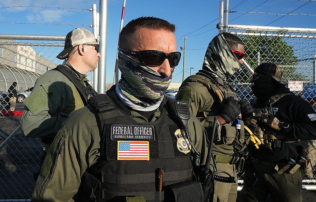 Two masked men and one without a mask, all in sunglasses stand in military gear outside a chain-link fence. The one in front has a badge reading "FEDERAL OFFICER Homeland Security."