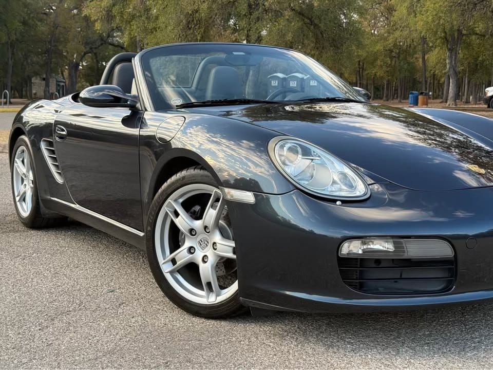 Front three-quarter view of a grey 2006 Porsche Boxster with the top down, parked on a tree-lined lot