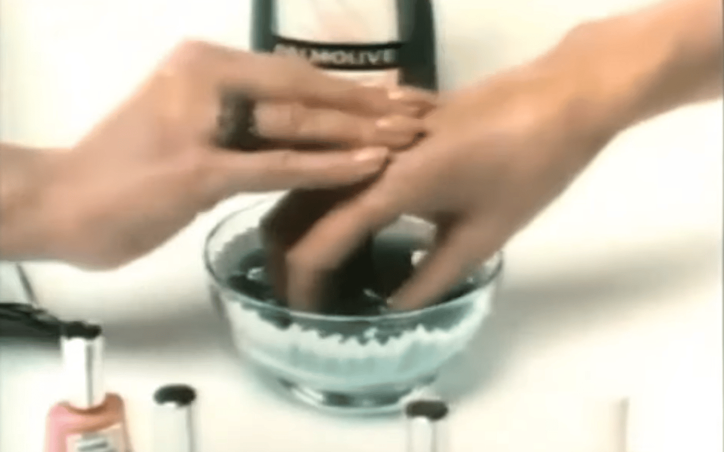 Two women's hands are visible near some bottles of nail polish. The fingers of the hand on the right are immersed in bowl of green liquid. The Palmolive logo is visible in the background. 