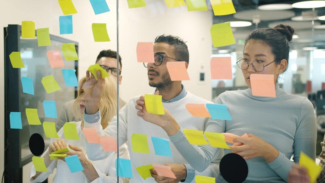 multi-ethnic team sticking colorful notes on glass board in office sharing ideas solving problems brainstorming concentrated on creative activity.