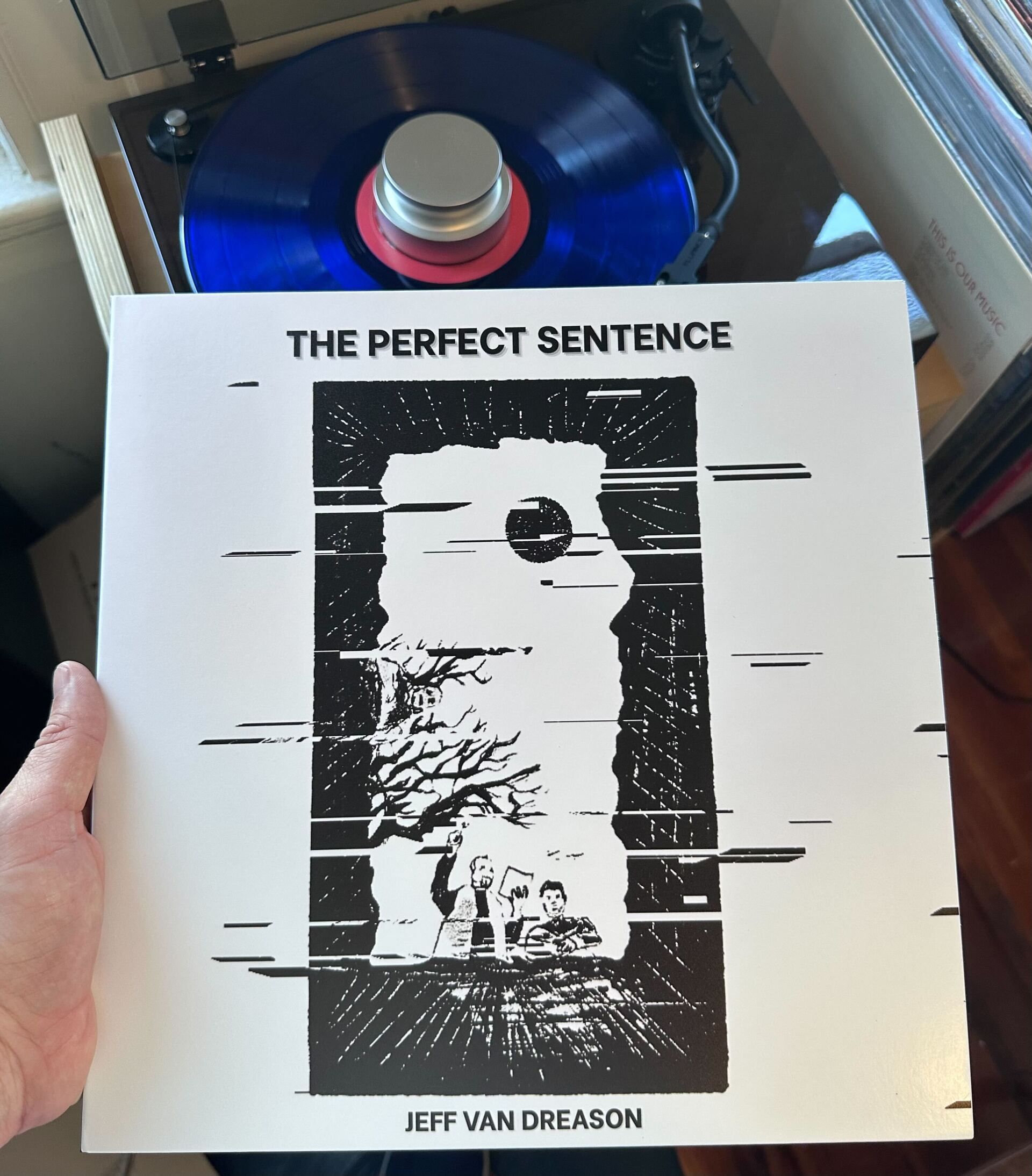Image shows the physical record cover of The Perfect Sentence, which shows people looking into a grave, with the point of view looking up at them. Behind te cover is the blue vinyl record on a record player, with a row of records organized beside it.