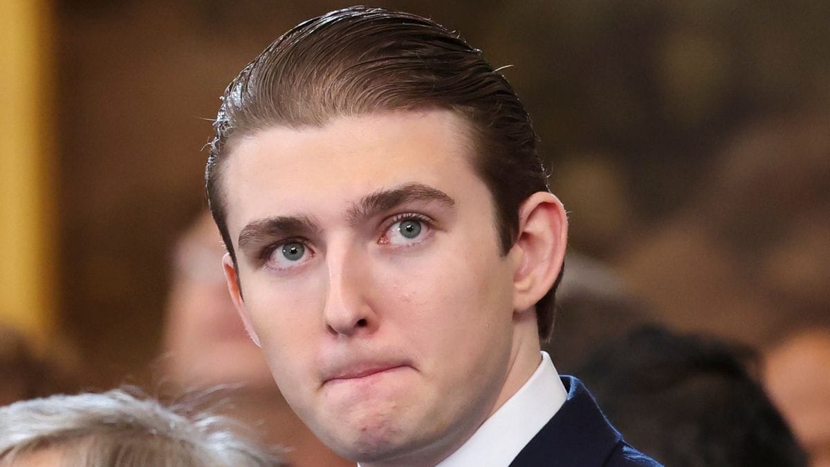 A white man wearing a dark suit looks to the right while standing indoors. People can faintly be seen in the background and left side of the image.