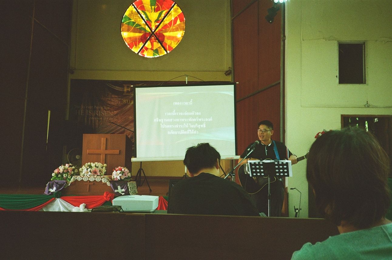 A thai pastor leads worship on guitar. Lyrics are projected on a screen.