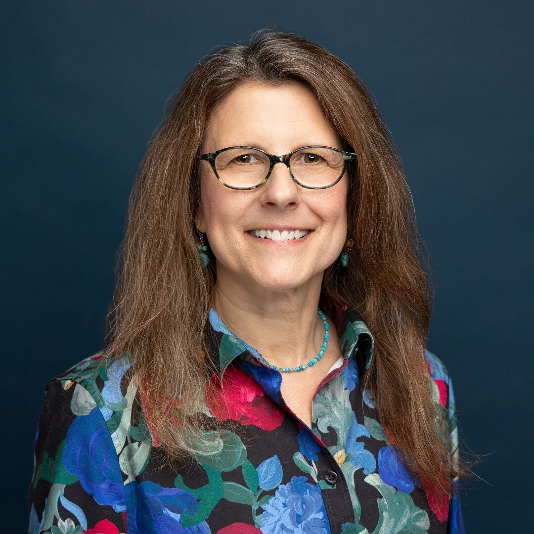 A woman with long brown hair and glasses smiles warmly against a dark blue background. She is wearing a colorful floral-patterned shirt and a turquoise beaded necklace with matching earrings.