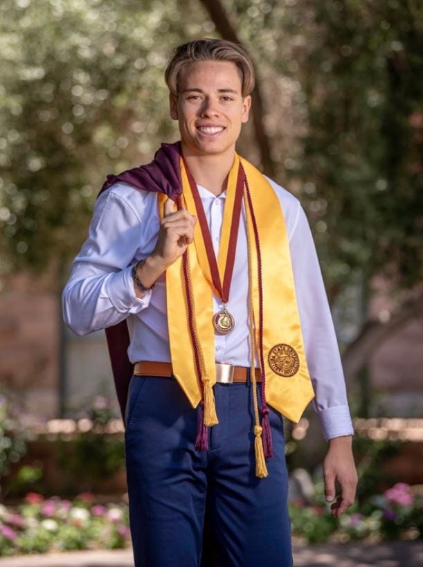 A proud graduate in a white shirt, blue pants, and yellow stole with a medal, smiling outdoors with a purple gown draped over the shoulder, surrounded by greenery and flowers.