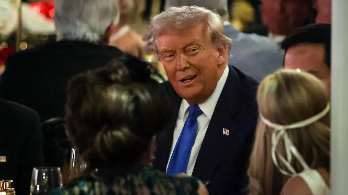A white man wearing a black suit grins as he talks to people sitting near him in a crowded room.