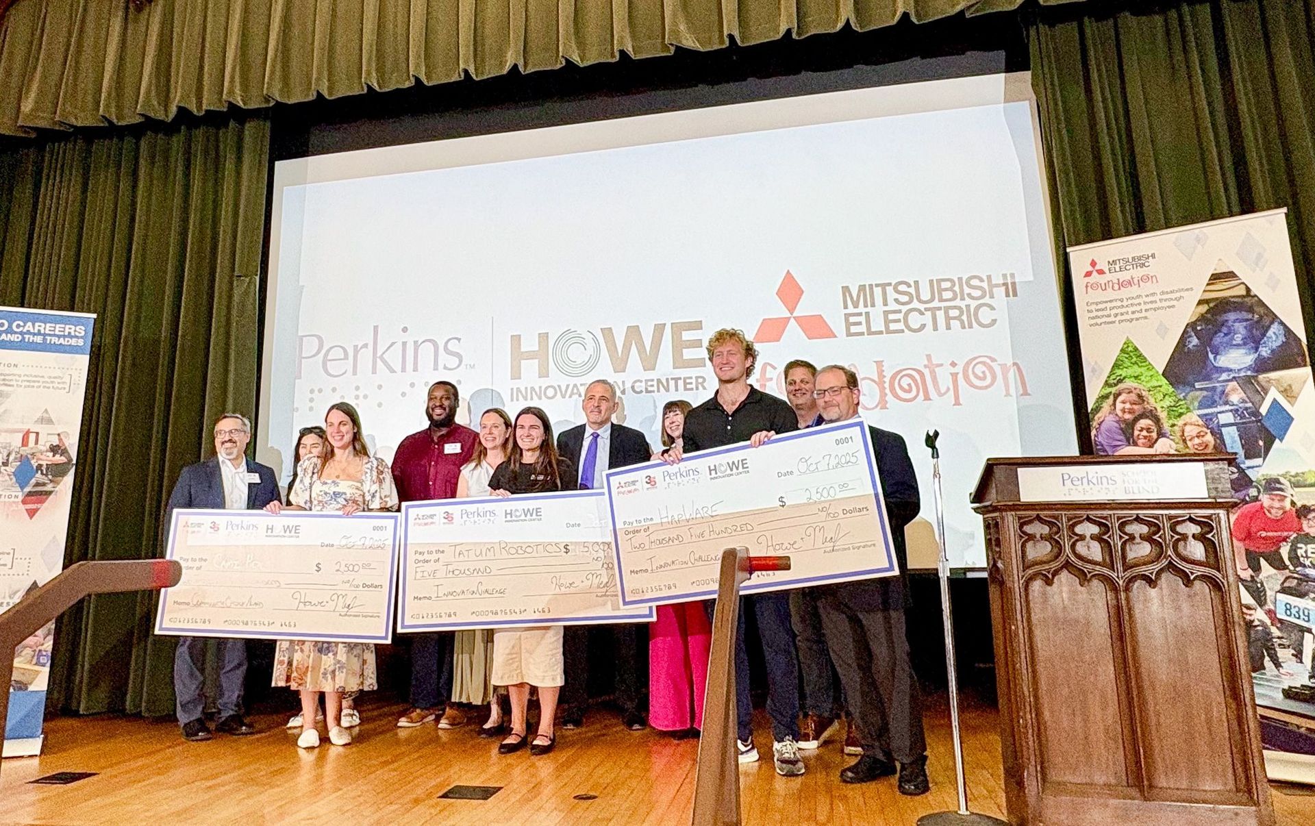 Jack Walters of HapWare stands on stage with other award recipients and presenters, holding an oversized $2,500 check made out to HapWare. The group smiles for a photo in front of a large projection screen showing the logos of Perkins School for the Blind, Howe Innovation Center, and Mitsubishi Electric Foundation.