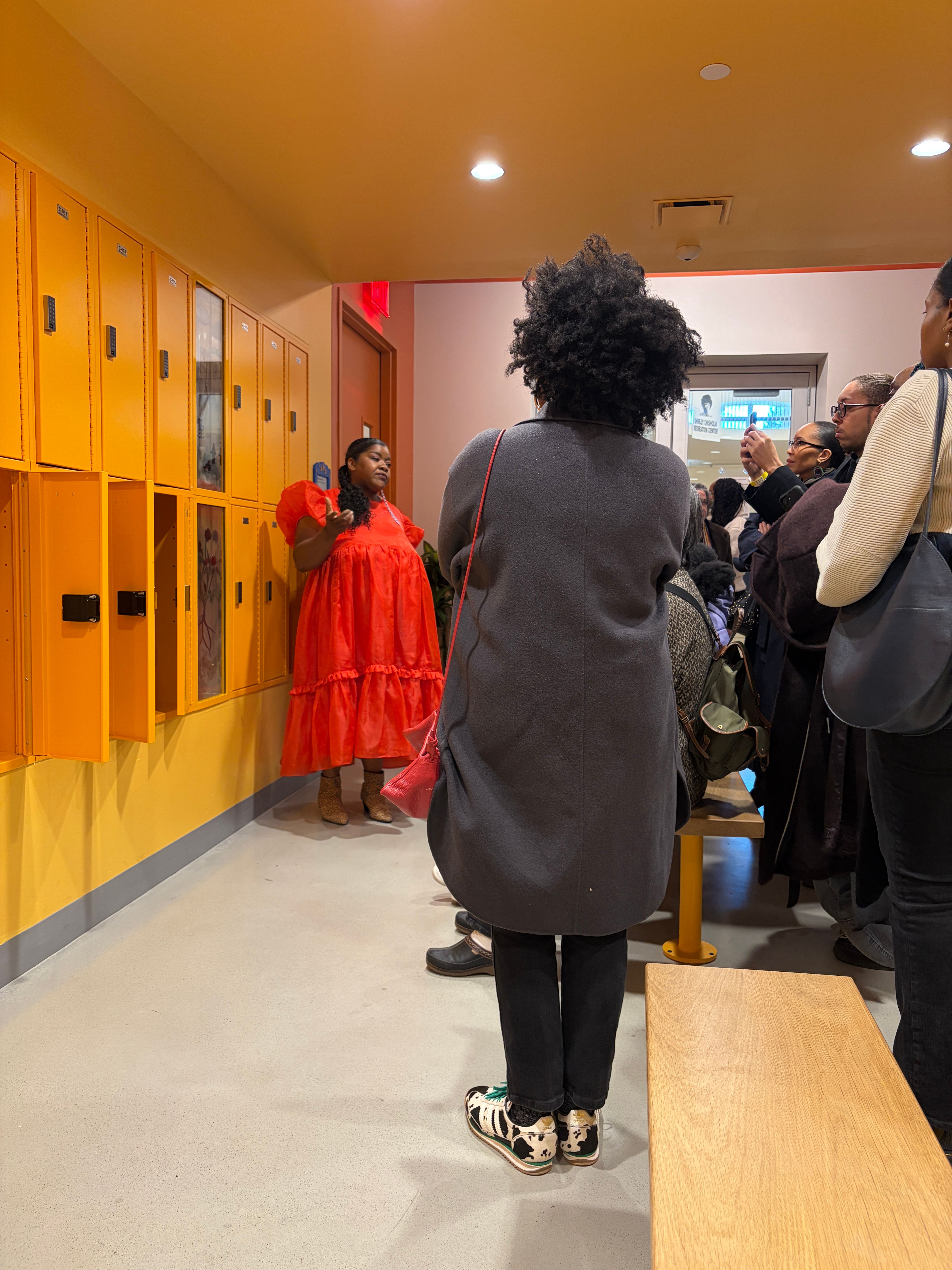 A Black woman in a red dress speaking to a crowd of people in a yellow locker room