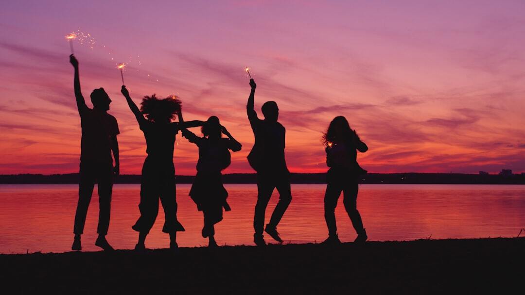 Group of friends men and women are dancing outdoors near lake with bengal lights having fun at night in darkness, beautiful colorful sky and water in background.
