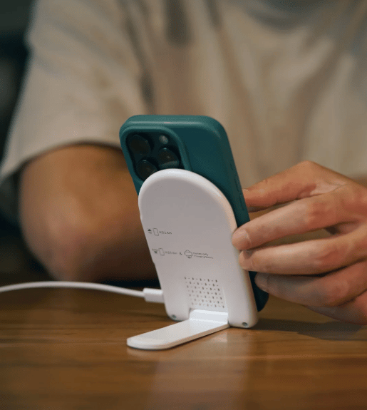 Person using a smartphone on a white wireless charging stand at a wooden desk, showing practical branded tech gifts for hybrid work setups.